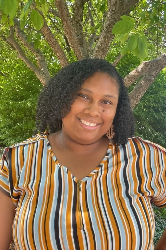 A licensed professional smiling outdoors with a tree and green leaves in the background, wearing a striped shirt and a leaf-shaped earring.