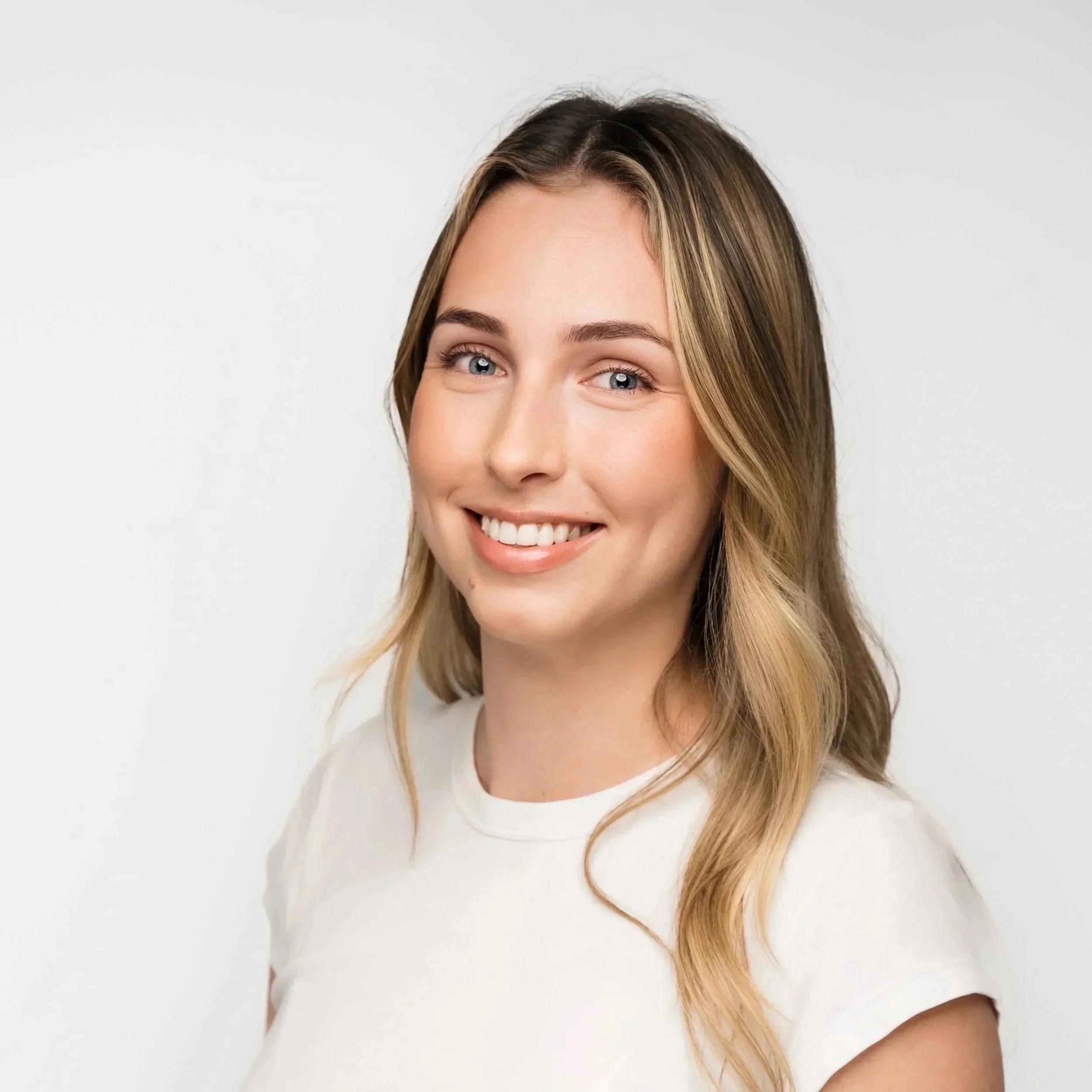 A smiling young woman with long, wavy blonde hair wearing a white t-shirt, standing against a plain white background.