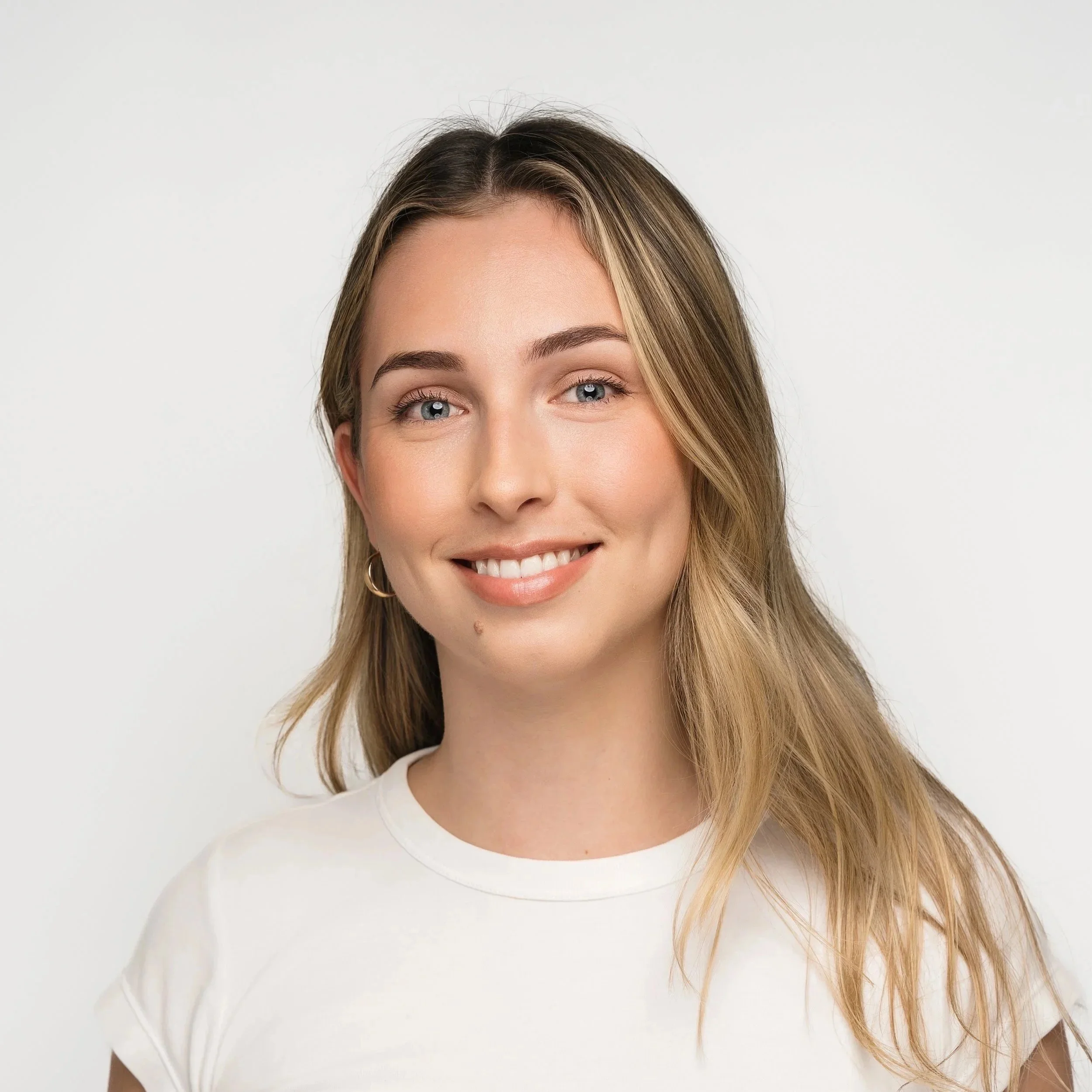 A young woman with long, light brown hair and blue eyes smiling at the camera, wearing a white t-shirt and small hoop earrings against a plain white background.