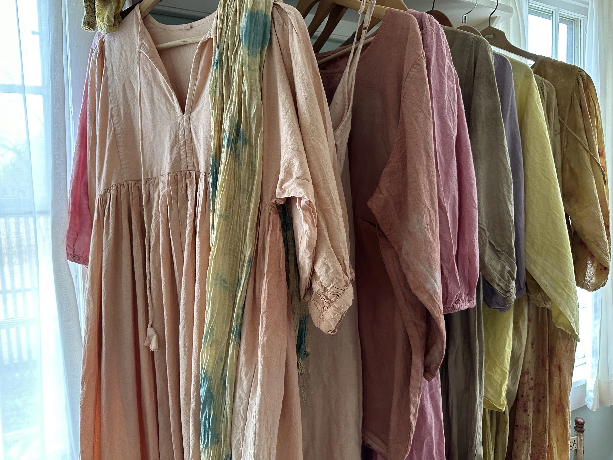 A collection of pastel-colored, loose-fitting linen dresses hanging on a clothing rack near a window.