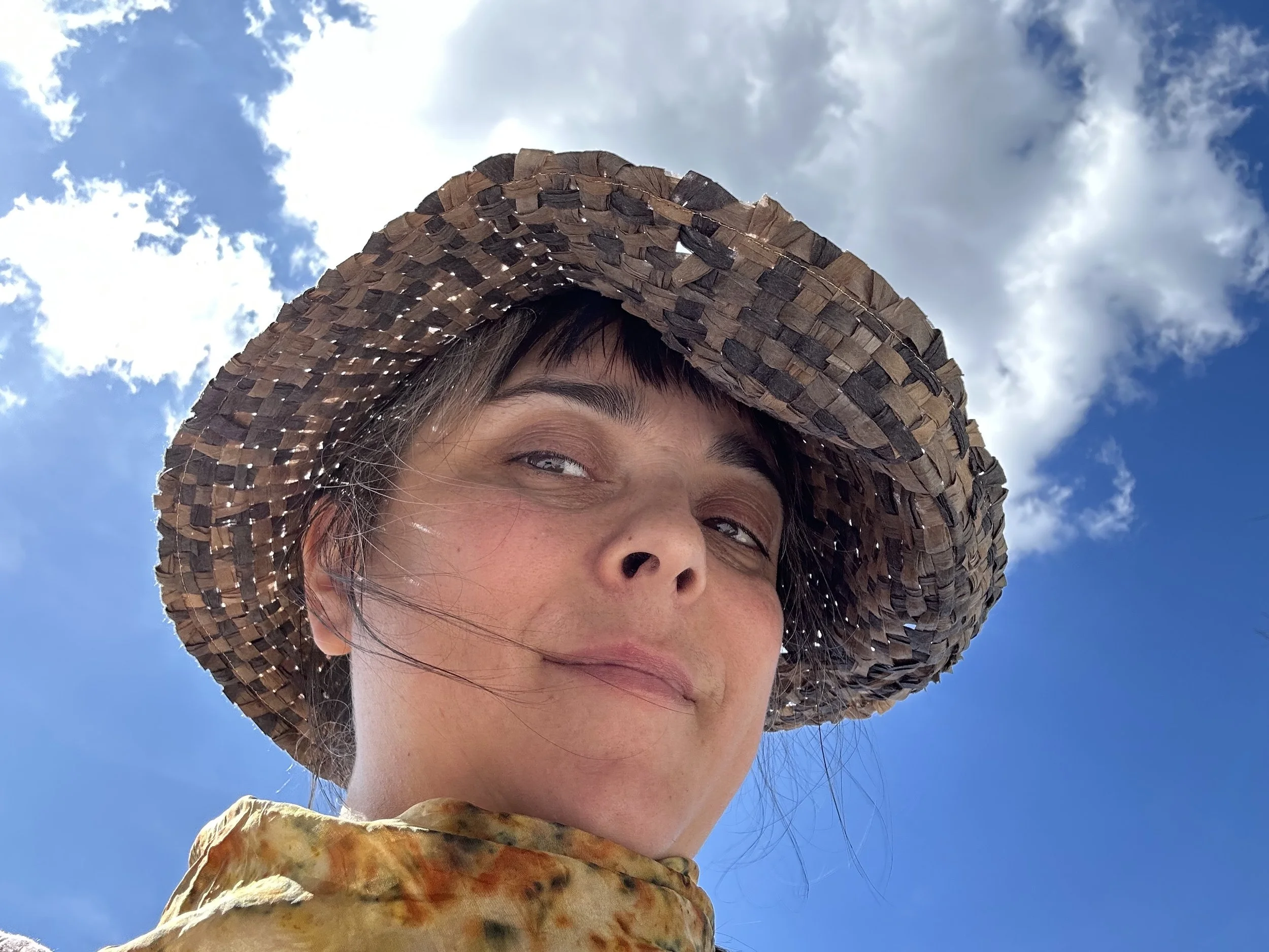 Close-up of a woman wearing a wide-brimmed woven straw hat outdoors against a blue sky with white clouds, smiling softly.