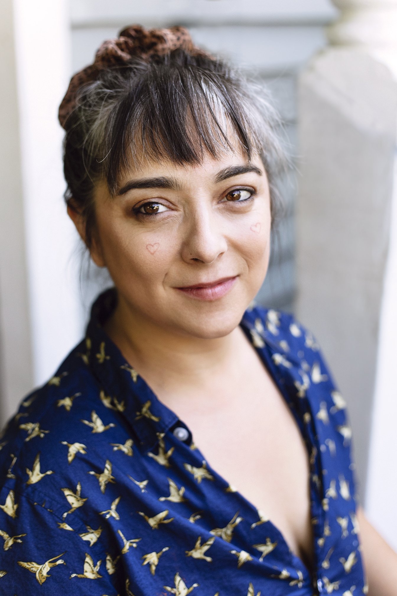 Close-up of a woman with short, tousled dark hair with lighter highlights, soft makeup with a touch of gray eyeshadow, small hearts drawn on her cheeks, wearing a navy shirt with a bird pattern, standing near a window with sunlight.