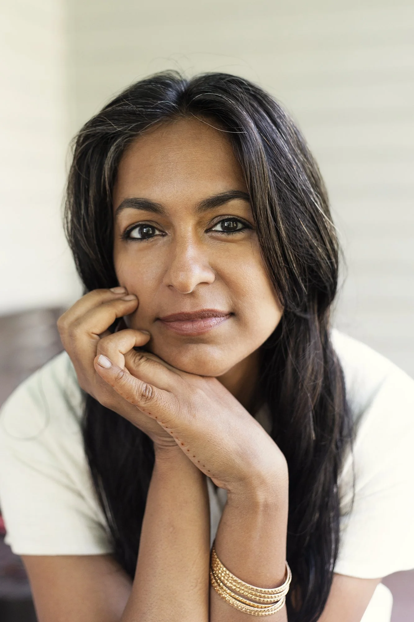 Close-up of a woman with long dark hair, looking at the camera with a slight smile, resting her face on her hands, wearing a white top and gold bracelets.