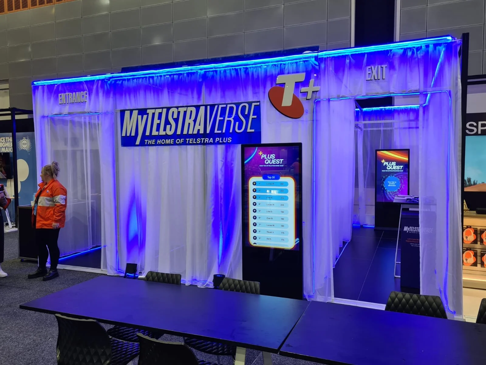 An exhibition booth for MY TELSTARVERSE, a Telstra Plus event, with blue neon lighting, white curtains, digital screens displaying leaderboard and promotion content, and a woman in an orange jacket standing nearby.