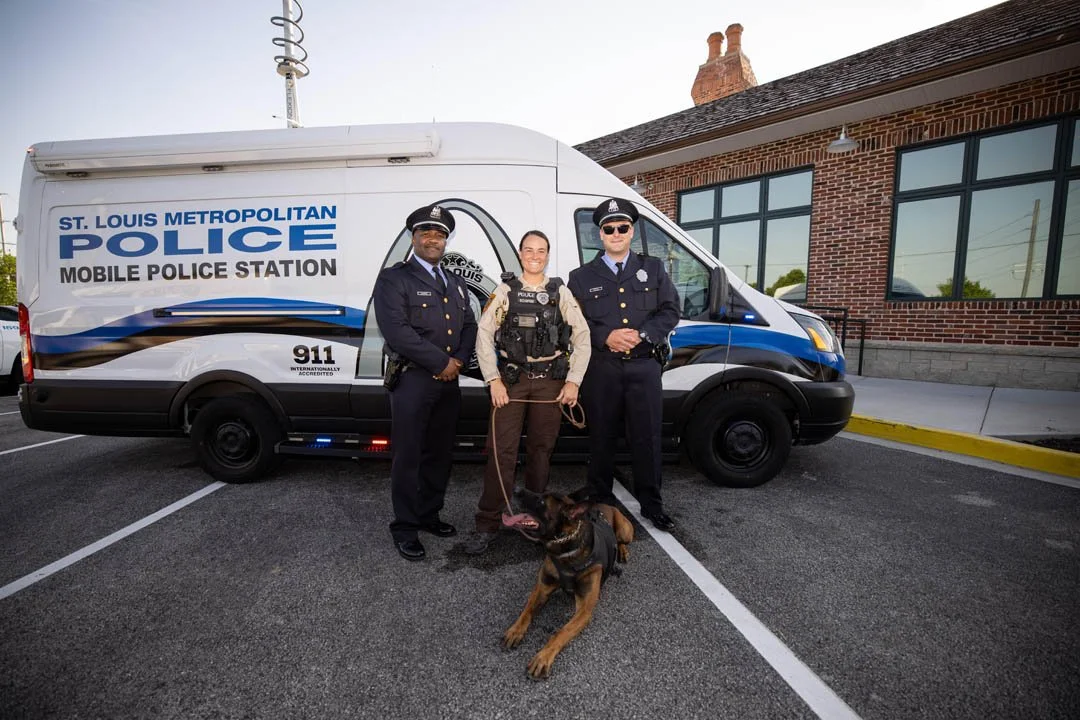 Three police officers standing in front of a marked police van, with a police dog on a leash, outside a brick building.