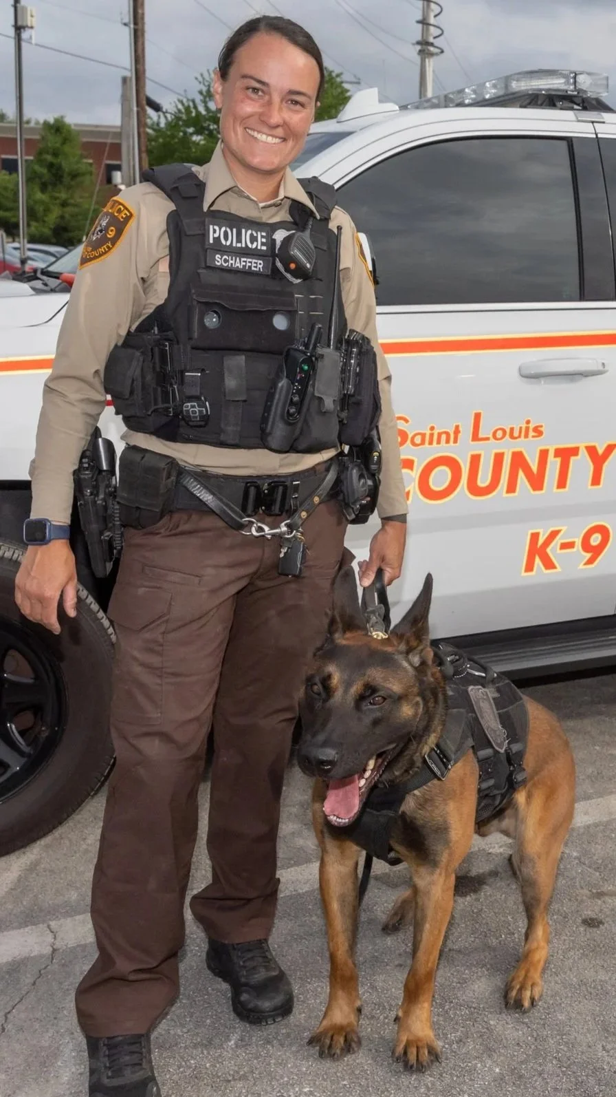 A female police officer standing next to her police dog in front of a Saint Louis County police vehicle. The officer is smiling and wearing a uniform with a tactical vest, and the dog is wearing a harness.