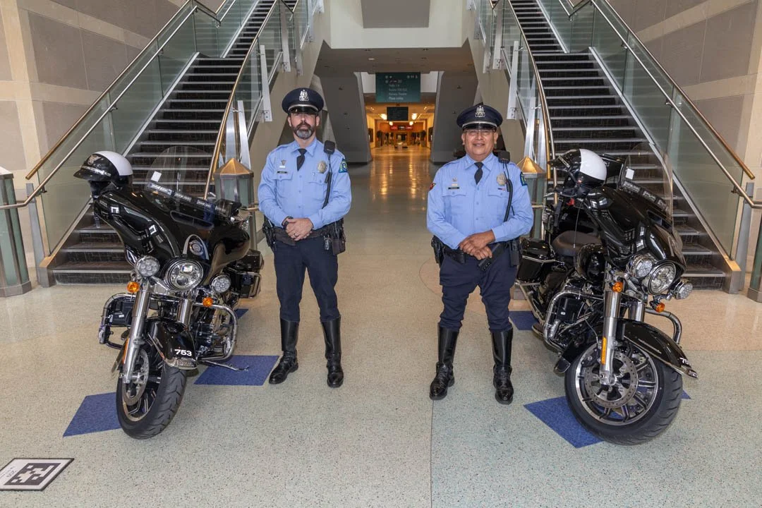 Two police officers standing in front of two black motorcycles inside a modern building with stairs on each side.
