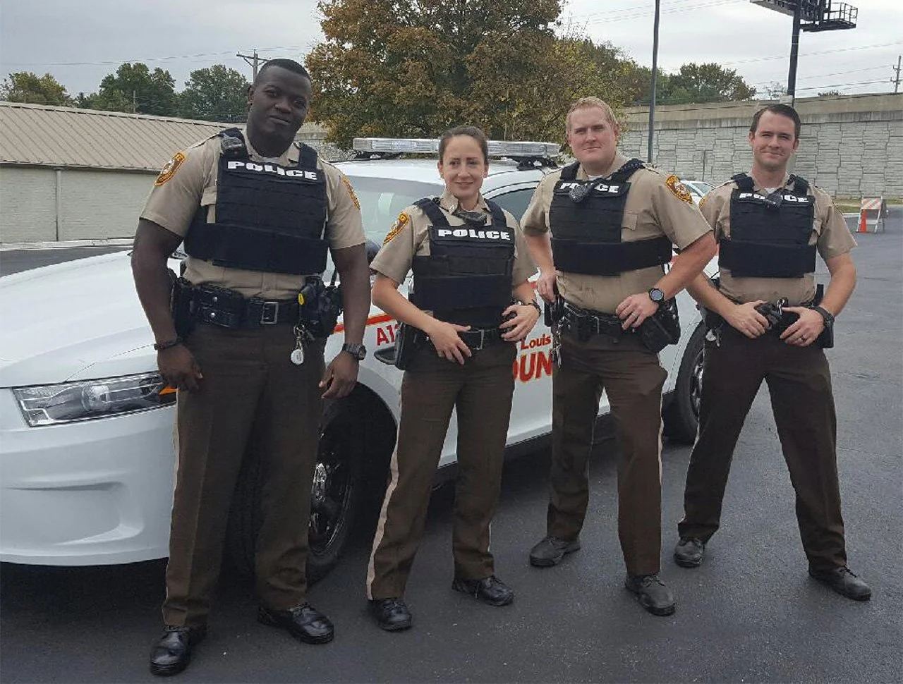 Four police officers standing in front of a police car outdoors during daytime. They are in uniform with bulletproof vests and have various equipment on their belts.