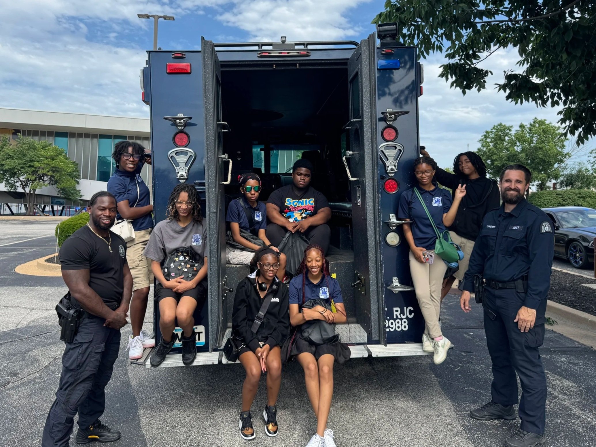 Group of people, including children and adults, posing in front of a police vehicle outdoors on a partly cloudy day.