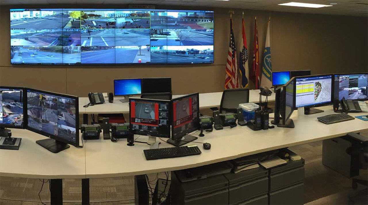 A security control room with multiple monitors displaying surveillance footage of parking lots and streets.