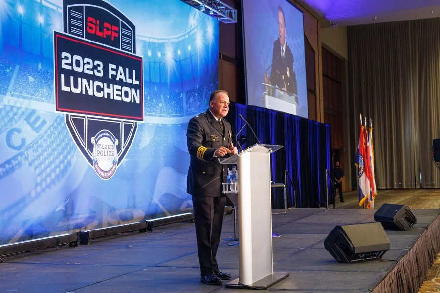 A man in a police uniform giving a speech at a podium during the 2023 Fall Luncheon hosted by the St. Louis Police Officers Association, with a large banner and a screen behind him, and flags on the right side of the stage.