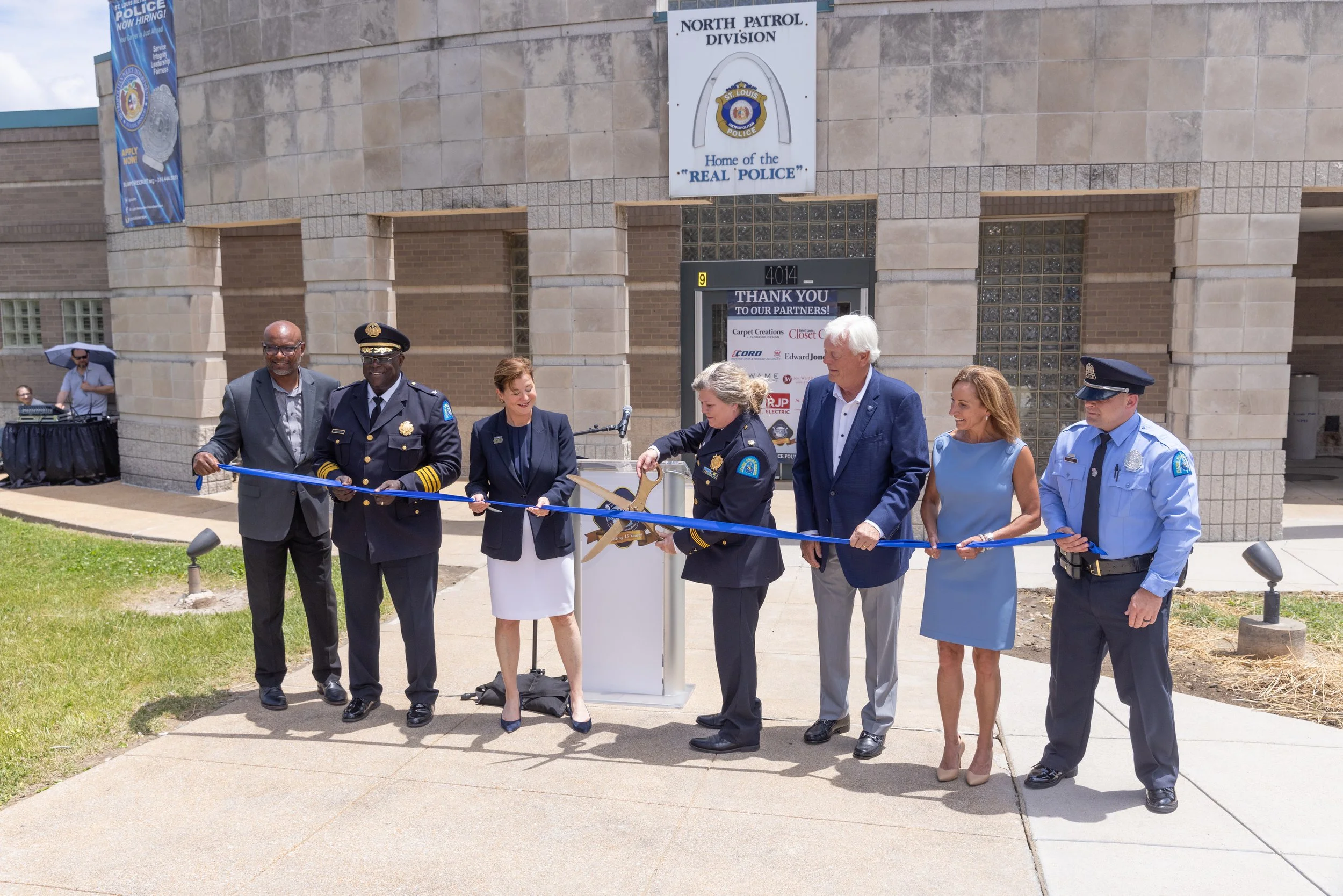 A group of seven people, including police officers and civilians, stand outside a beige brick building with a sign reading "North Patrol Division, Home of the 'Real Police'". They are participating in a ribbon-cutting ceremony for a police division, with one person cutting the ribbon and others observing and smiling. A man in a blue suit and a woman in a blue dress hold the ribbon, while a police officer in uniform and a woman in a blazer also assist.