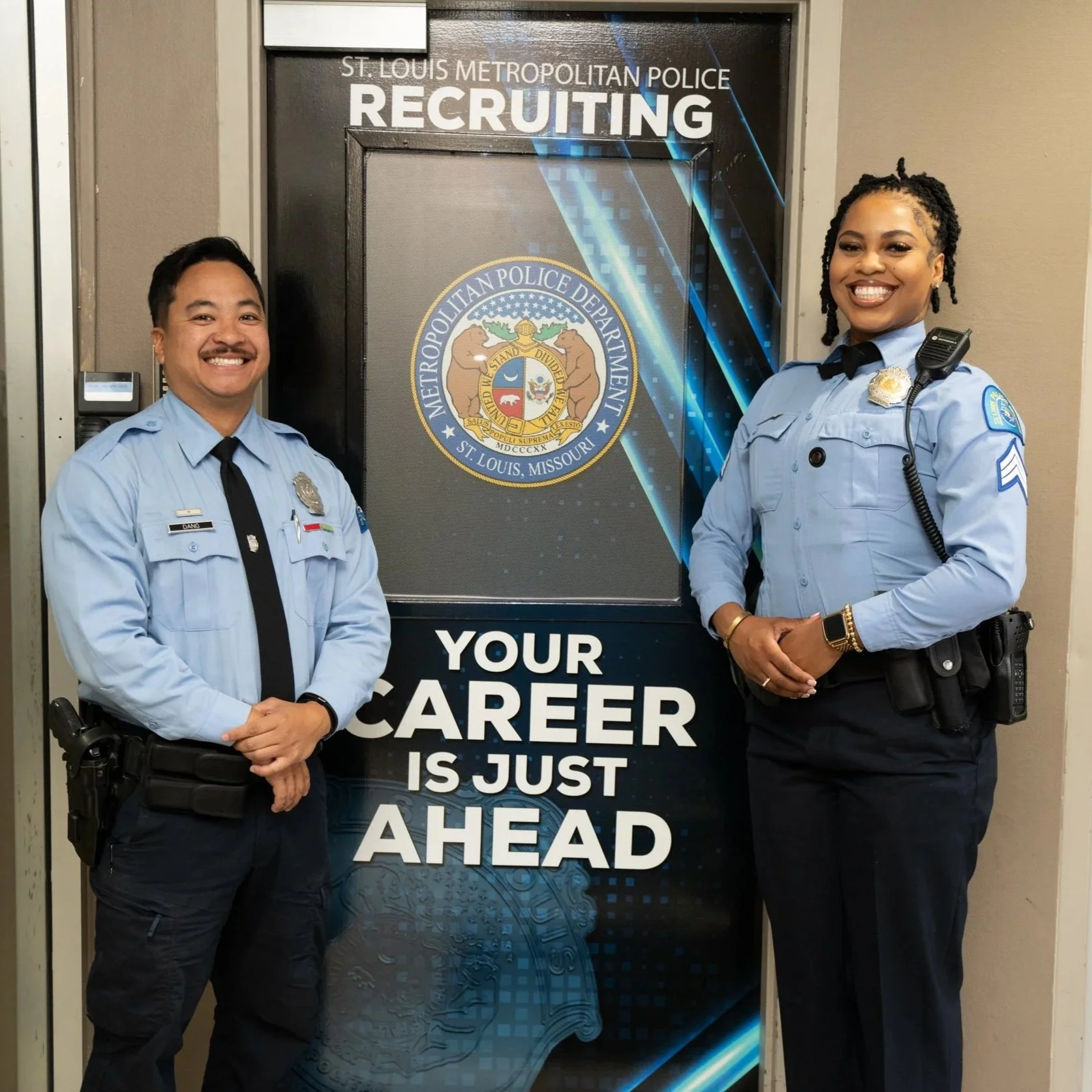 Two police officers dressed in light blue uniforms smiling and standing in front of a black recruitment sign with the seal of the St. Louis Metropolitan Police Department. The sign features text about career opportunities, and the officers are smiling with their hands clasped.