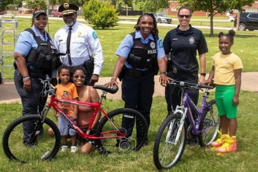 Group of three police officers, two women and one man, standing outdoors with two children and two bicycles, in a park with trees and grass.