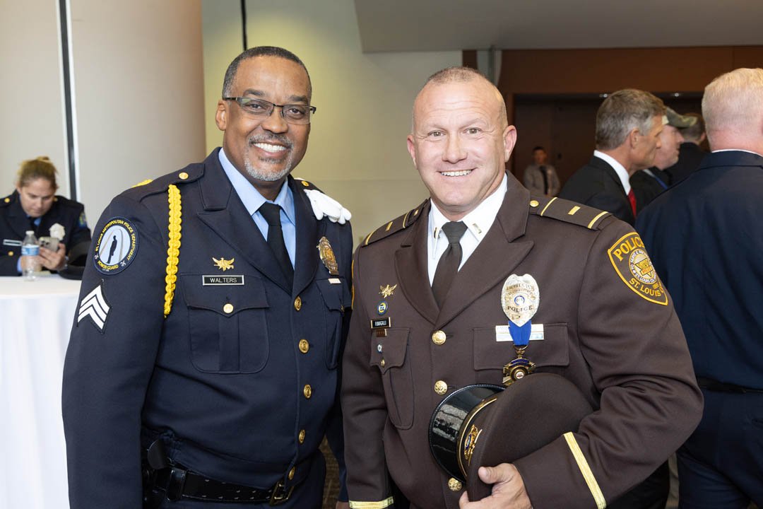 Two police officers in uniform smiling at a formal event, with other people in matching uniforms in the background.