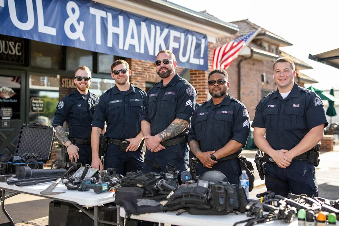 Five police officers in uniform standing behind a table with equipment, outdoors in front of a building with a blue banner reading 'THANKFUL & GRATEFUL!' and an American flag.