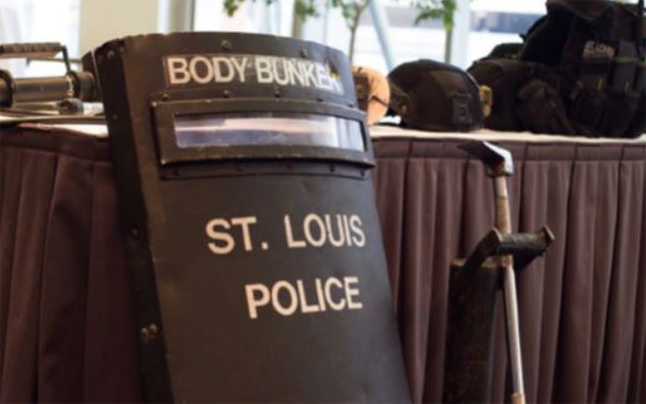 A police riot shield labeled "St. Louis Police" and a crutch are positioned against a table with bags and equipment on it.