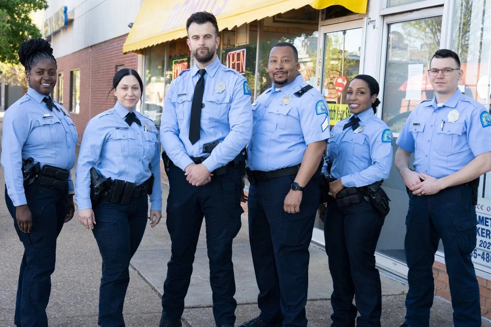 A diverse group of six police officers standing outside a storefront, smiling and posing for the camera.