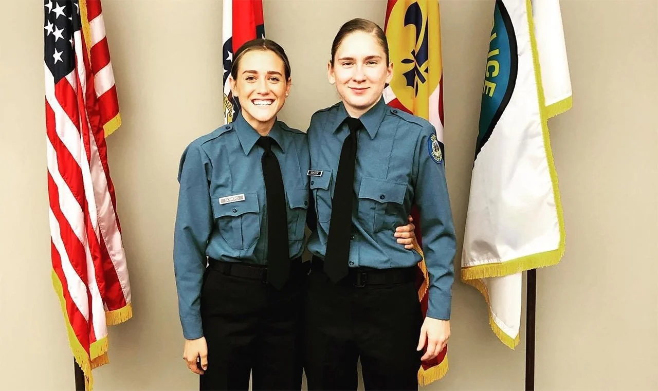 Two young women in police uniforms standing together in front of a row of American flags, smiling.