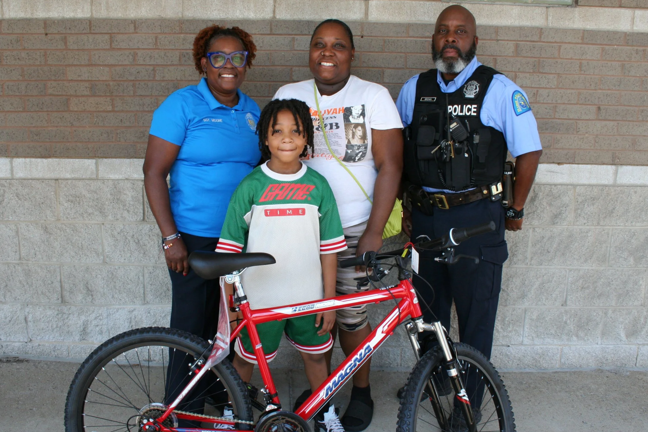 A group of four people, including a young girl with braids, a woman in a white T-shirt, a woman in a blue police uniform, and a man in a police uniform, standing against a brick wall. The young girl is holding a red bicycle with a black seat.