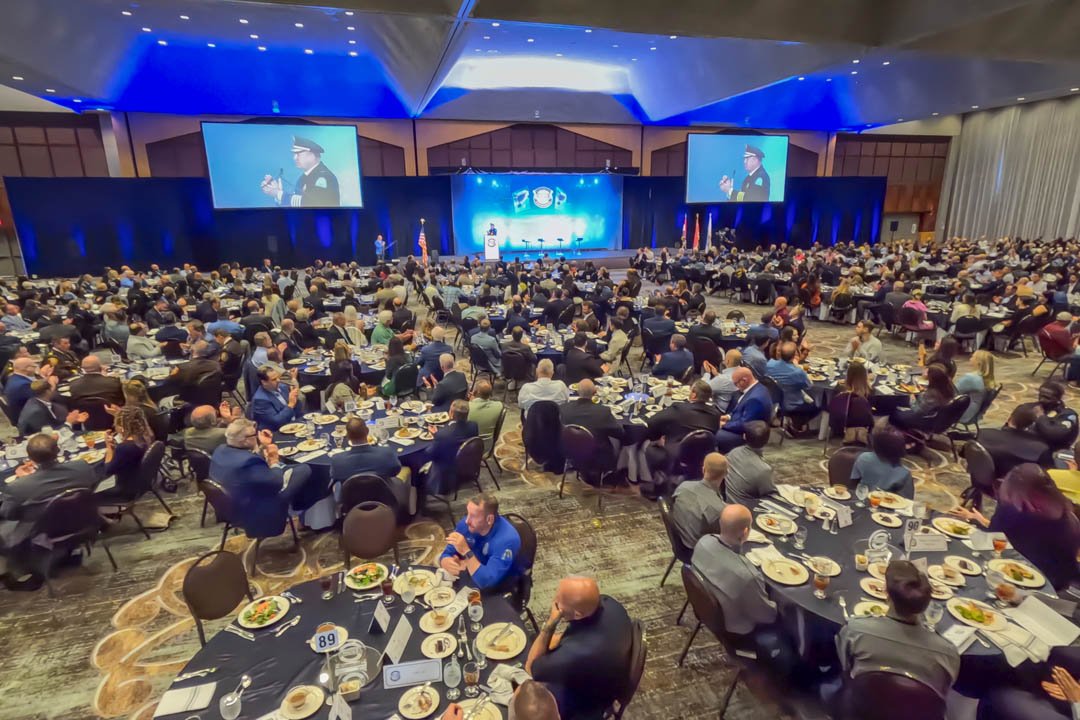 Large conference hall filled with round tables and people attending an event, with two large screens displaying a speaker in a uniform, and a stage with a podium and flags.