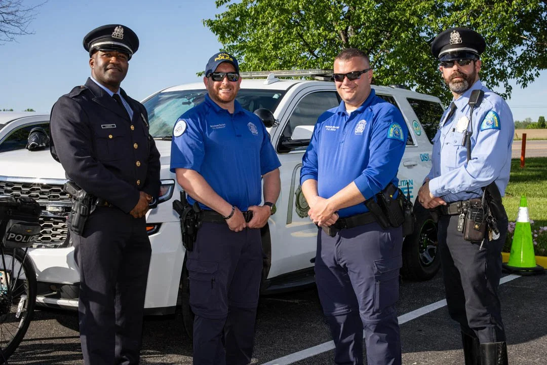 Four police officers standing next to a white police SUV, outdoors on a sunny day. Two officers are in dark uniforms, and two are in blue uniforms, all wearing sunglasses.