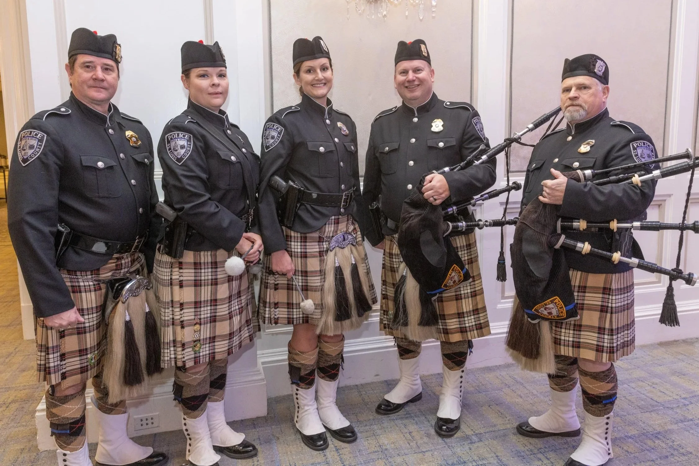 Group of five police officers dressed in traditional Scottish attire, including kilts, posing indoors against a light-colored wall with decorative molding. One officer is holding bagpipes.