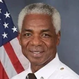 Portrait of a man with gray hair in uniform, standing in front of an American flag.