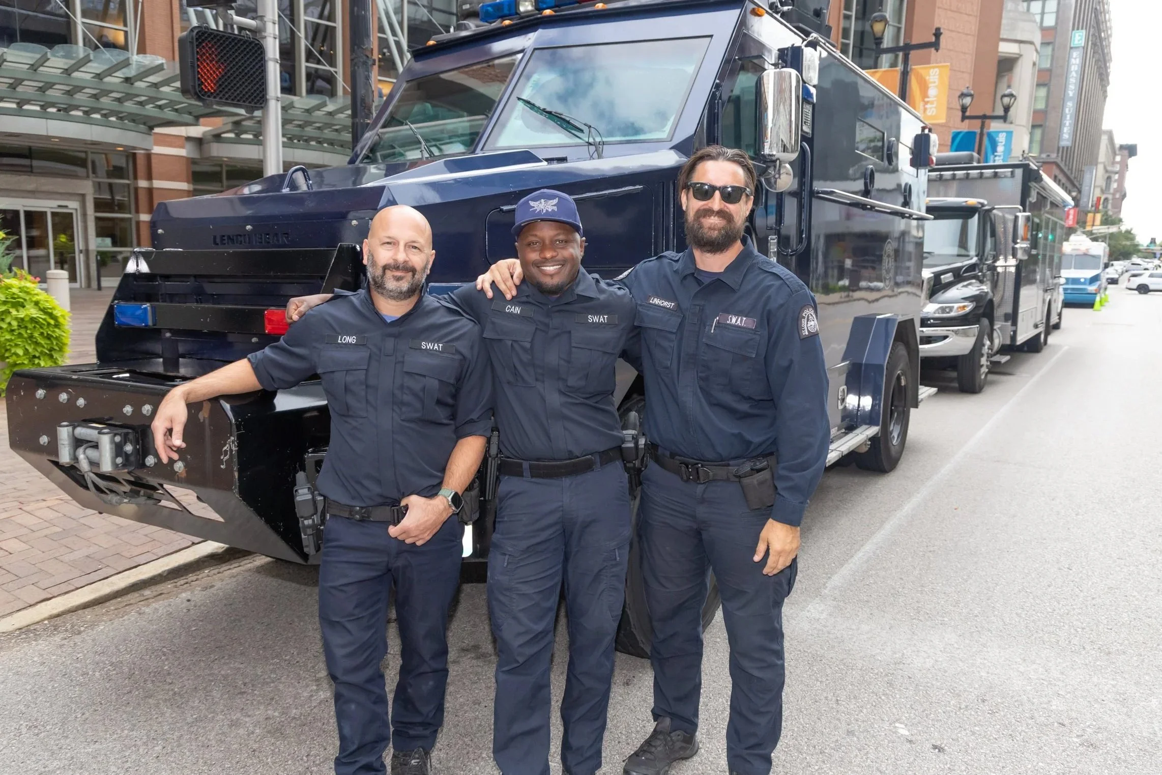 Three law enforcement officers in navy blue uniforms standing on the street smiling and posing for a photo in front of a police vehicle, with city buildings and traffic in the background.