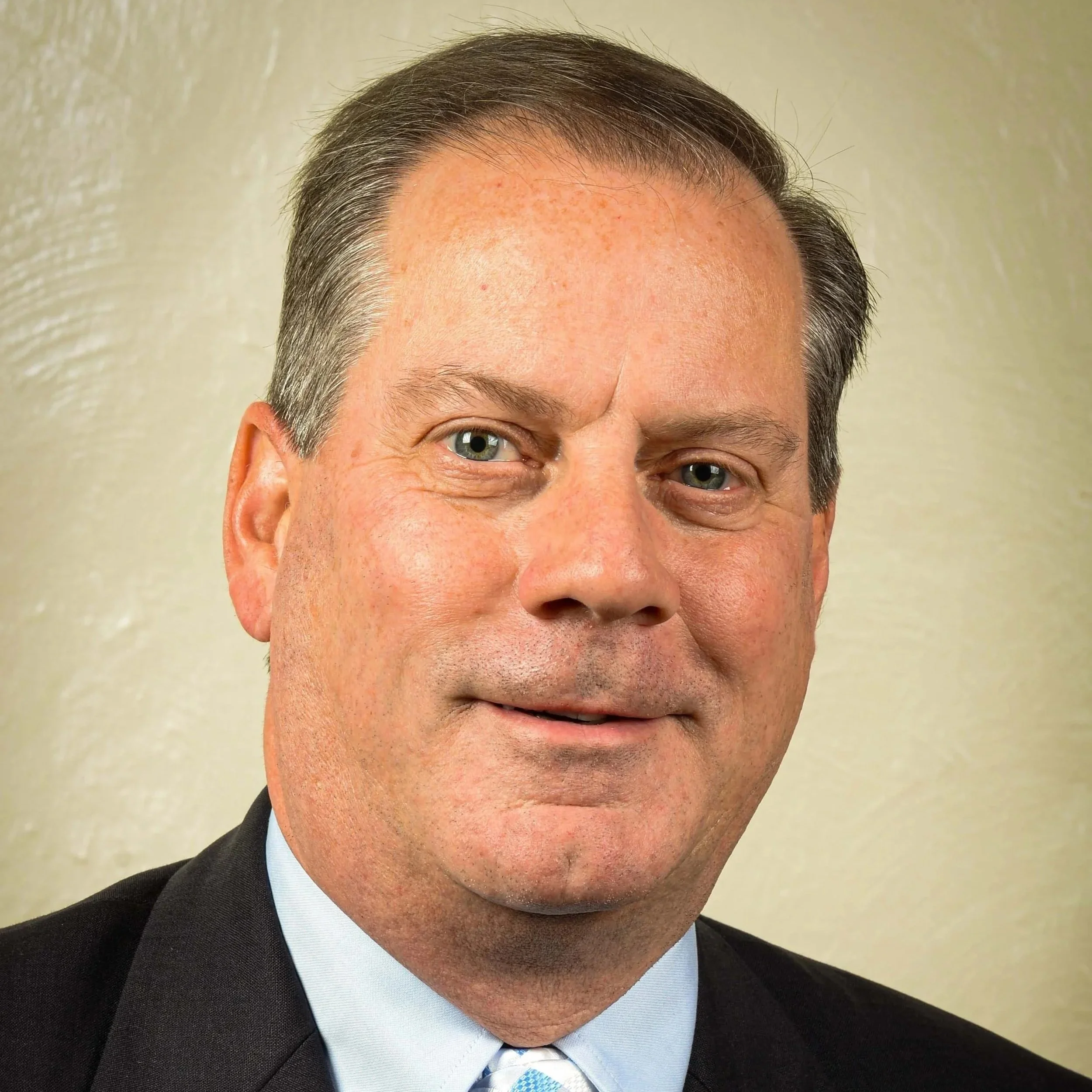 Close-up of a man with gray and brown hair, blue eyes, wearing a black suit jacket, white shirt, blue patterned tie, in front of a beige wall.