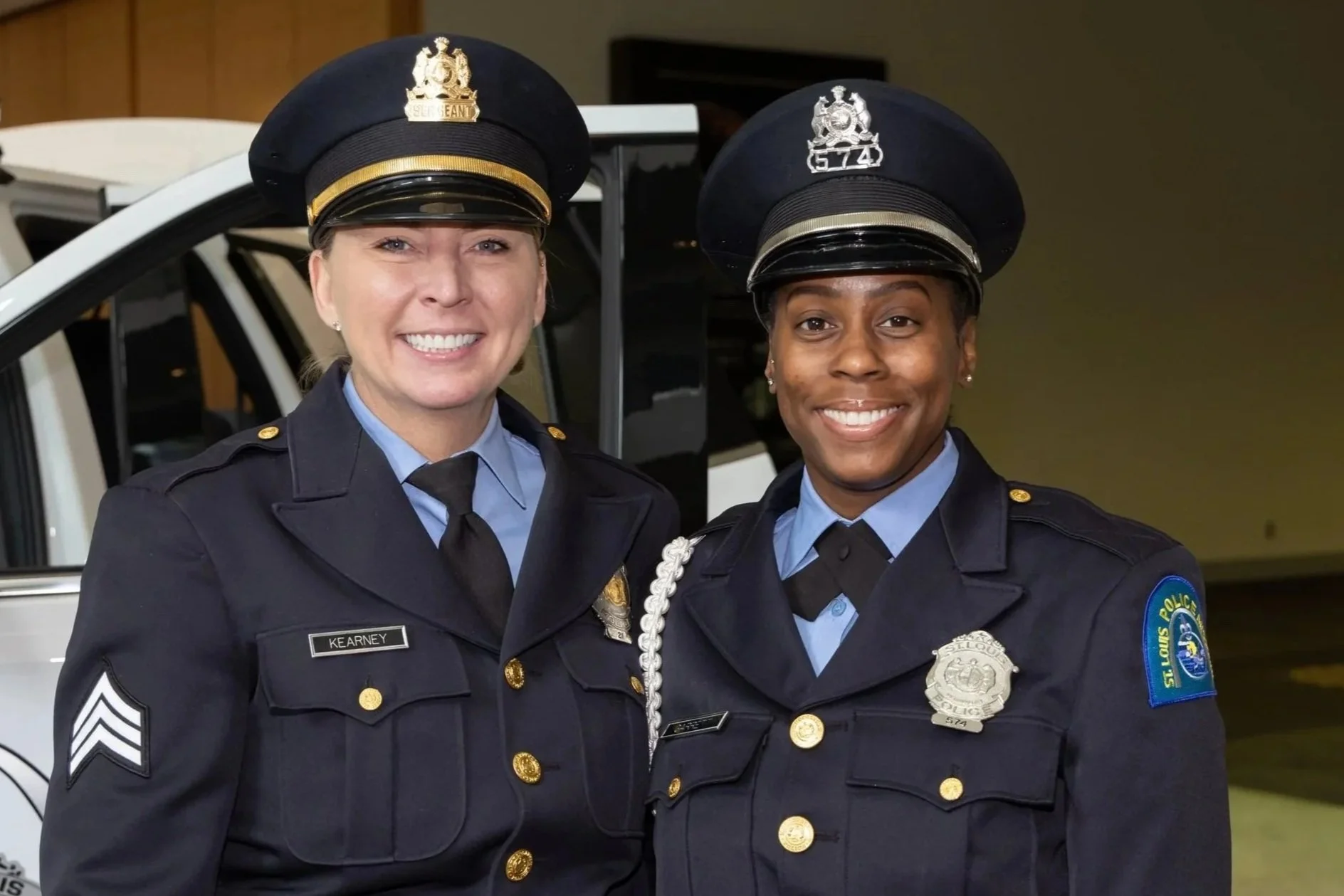 Two police officers in uniform smiling, one female on the left and one male on the right, standing in front of a police vehicle.