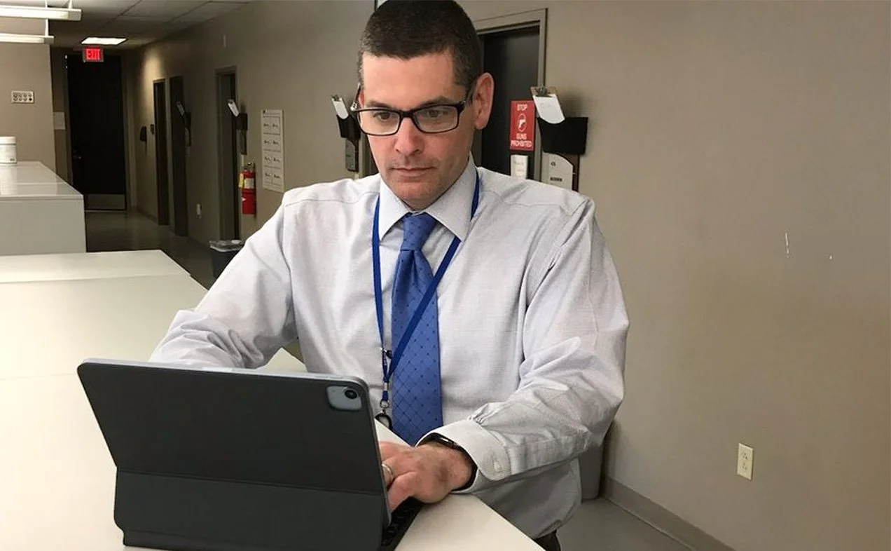 A man wearing glasses, a white dress shirt, and a blue tie is sitting at a desk, using a tablet device in a professional setting.
