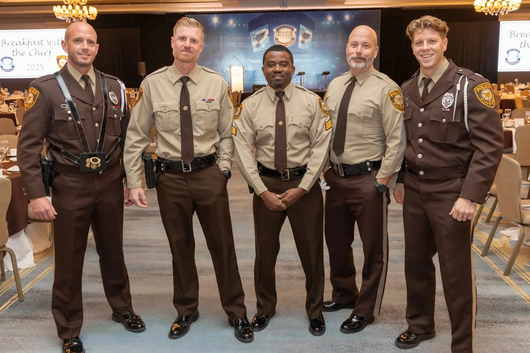 Group of five police officers standing together in a banquet hall, with tables and chairs visible in the background.