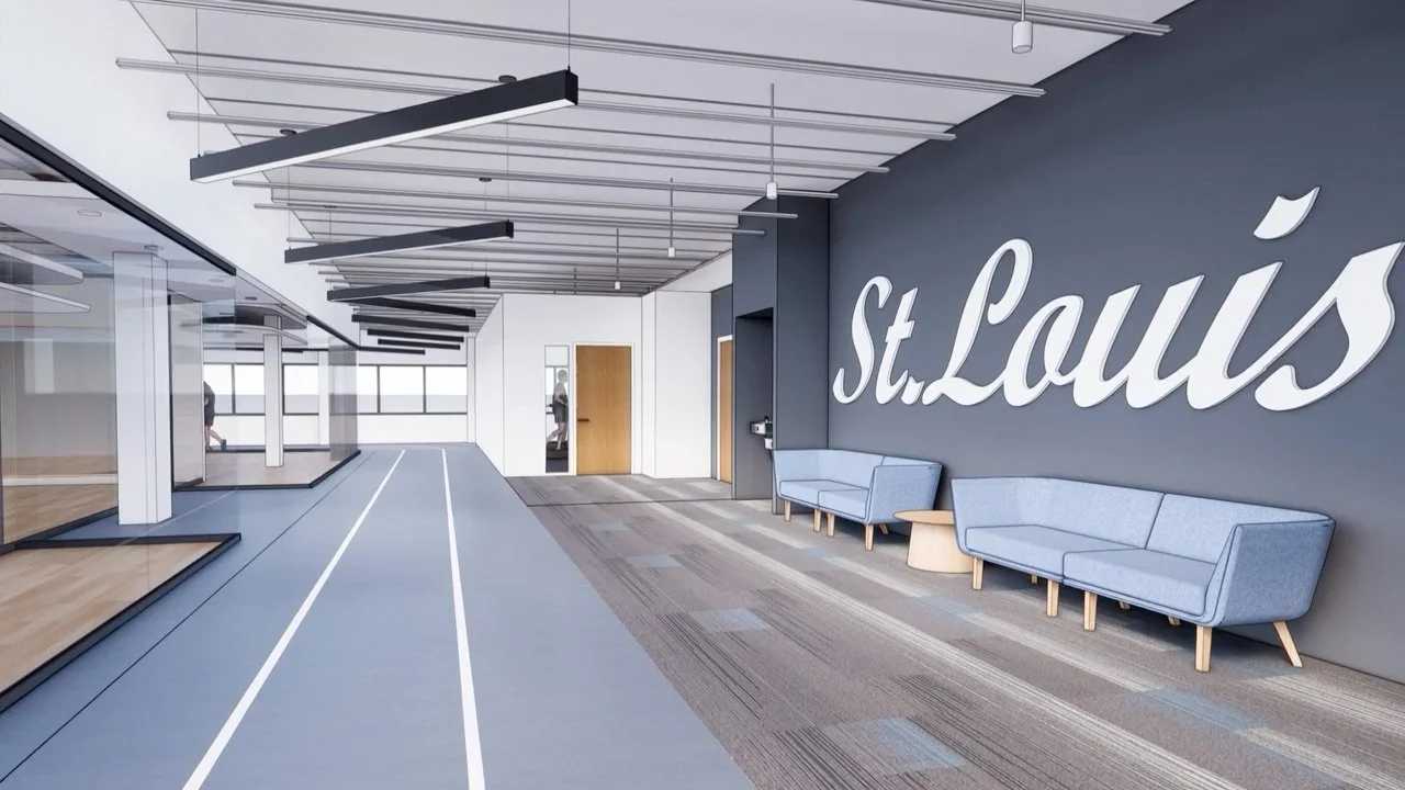 A modern indoor athletic training facility with a running track on the left and a waiting lounge on the right, featuring blue couches and a large wall sign that reads "St. Louis."