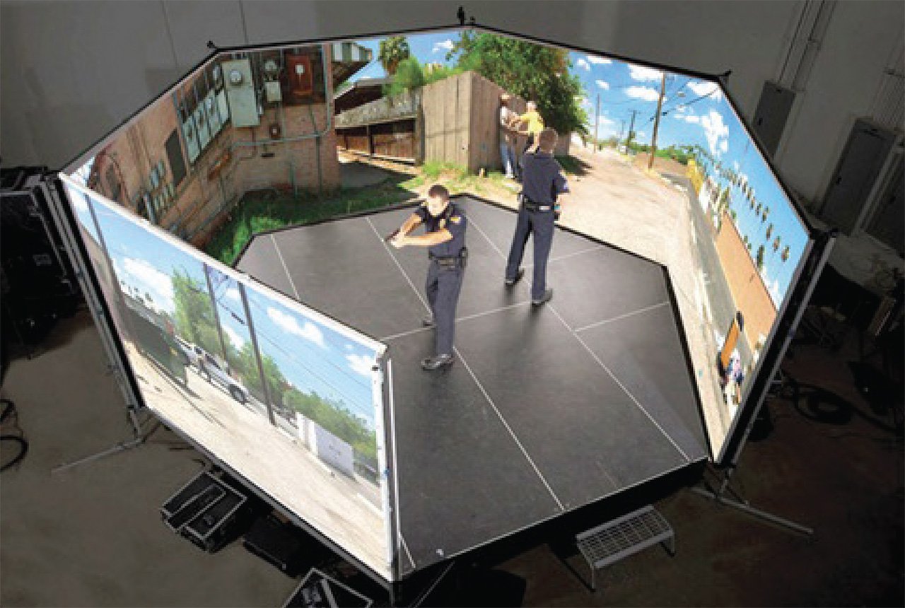 Two police officers setting up a large enclosed projection screen displaying an outdoor scene with a dirt road, wooden fence, and houses, inside a room.