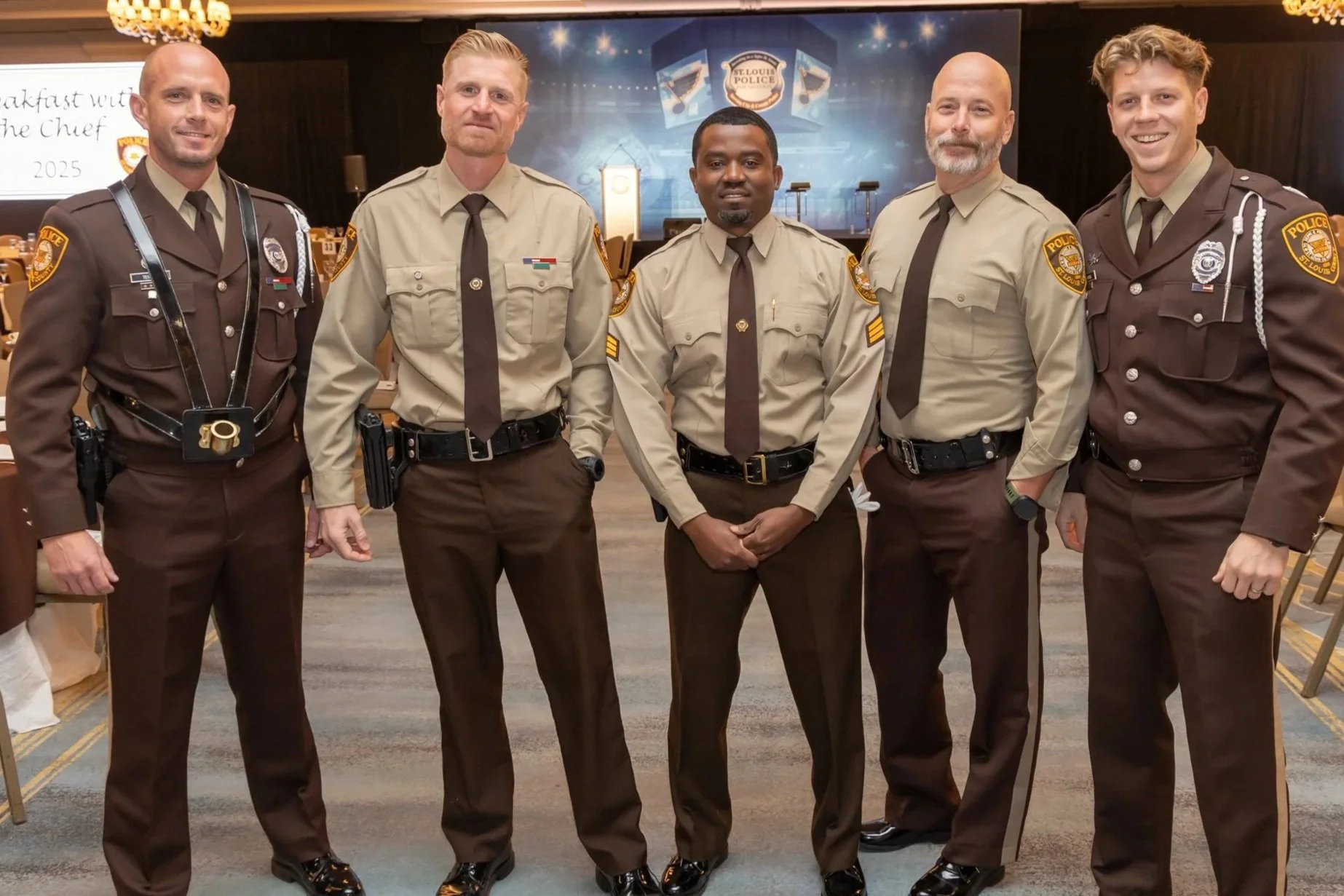 Five male police officers standing together indoors at a formal event, five are wearing tan uniforms and two are in dark brown uniforms with police patches on their sleeves, in front of a backdrop with police insignia and stage.