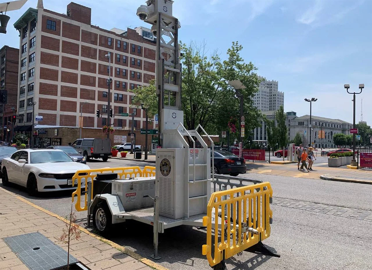 A street scene with a mobile surveillance or electrical unit on a trailer, yellow barricades, cars parked along the street, a building with a brick facade, trees, and pedestrians crossing the street in an urban area under a clear sky.