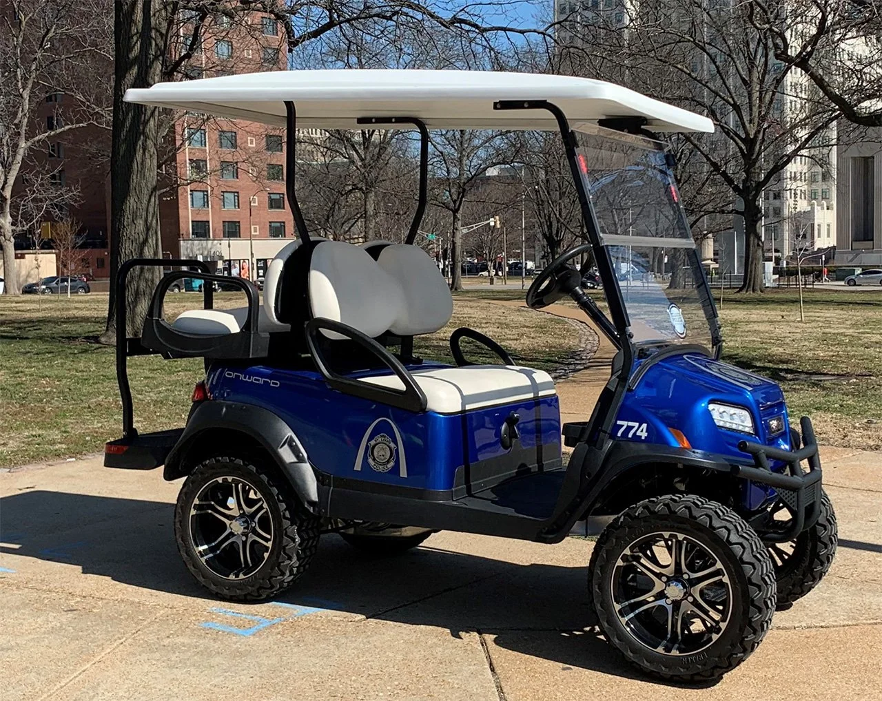 A blue police golf cart with a white roof parked on a sidewalk in a park, with trees and buildings in the background.