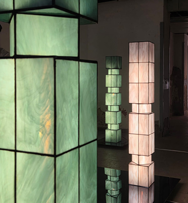 Three illuminated paper lanterns or light sculptures with geometric shapes, one green and two white, on a dark reflective surface in an indoor setting.