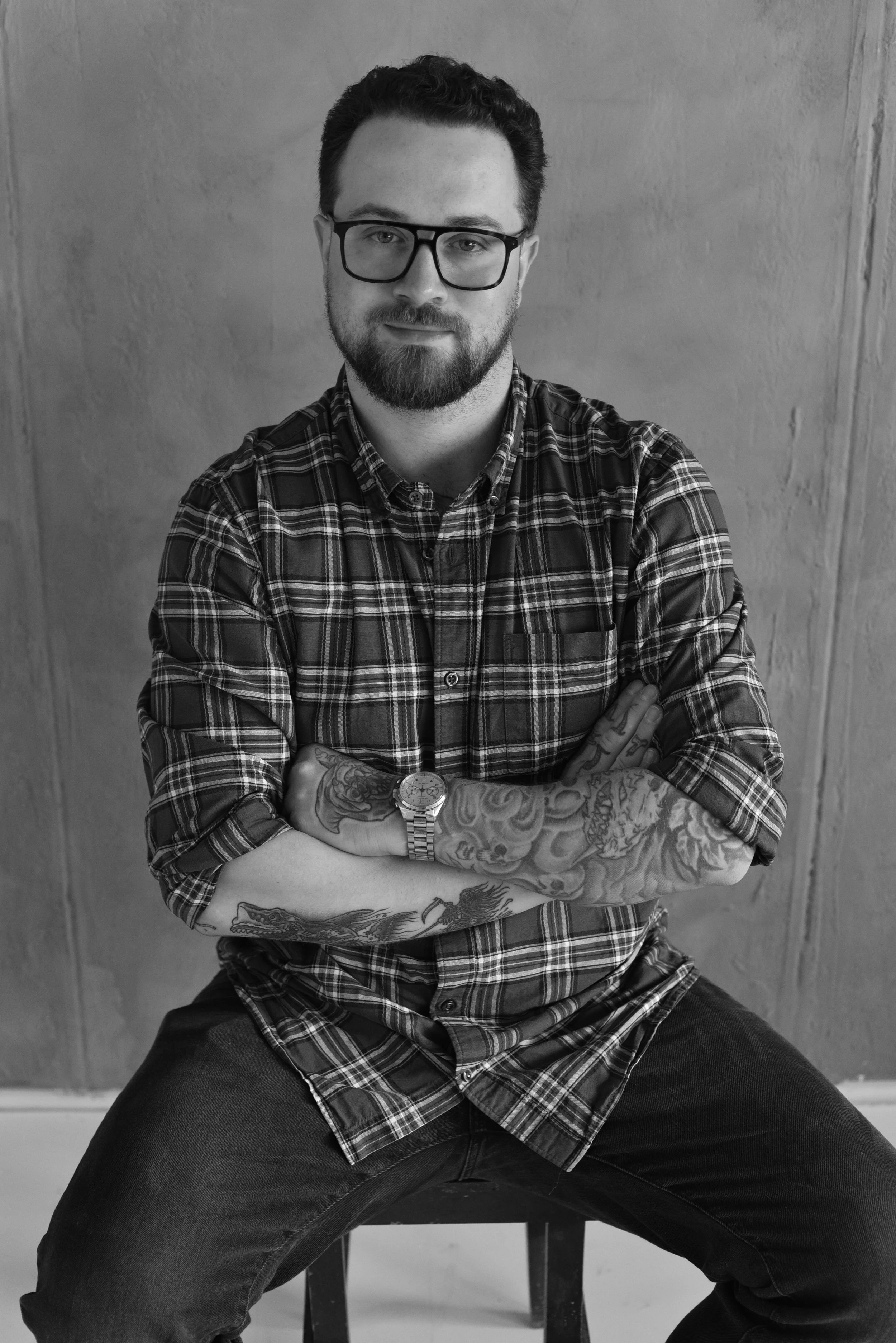 A man with glasses and a beard, wearing a plaid shirt with rolled-up sleeves, tattoos on his arms, and a wristwatch, seated on a stool with arms crossed against a plain background.