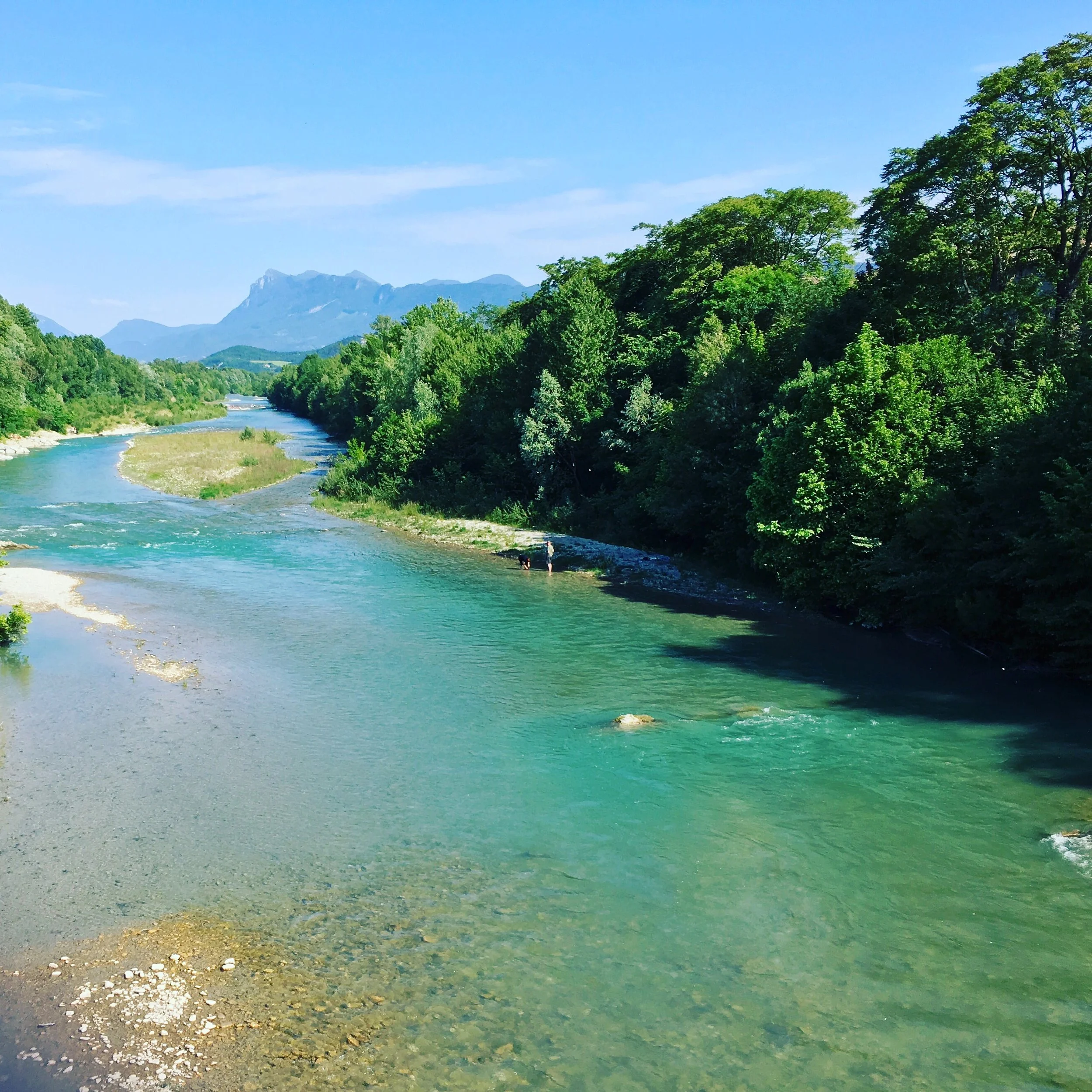 Vue panoramique d'une rivière turquoise entourée de forêts vertes luxuriantes, avec des montagnes en arrière-plan et un ciel bleu clair.