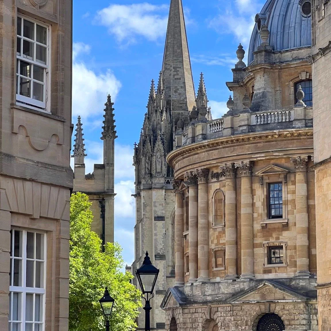 Vue d'une église gothique en pierre avec des tours et un dôme, entourée d'arbres verts et de lampadaires en fer, sous un ciel bleu avec quelques nuages, à Oxford en Angleterre.