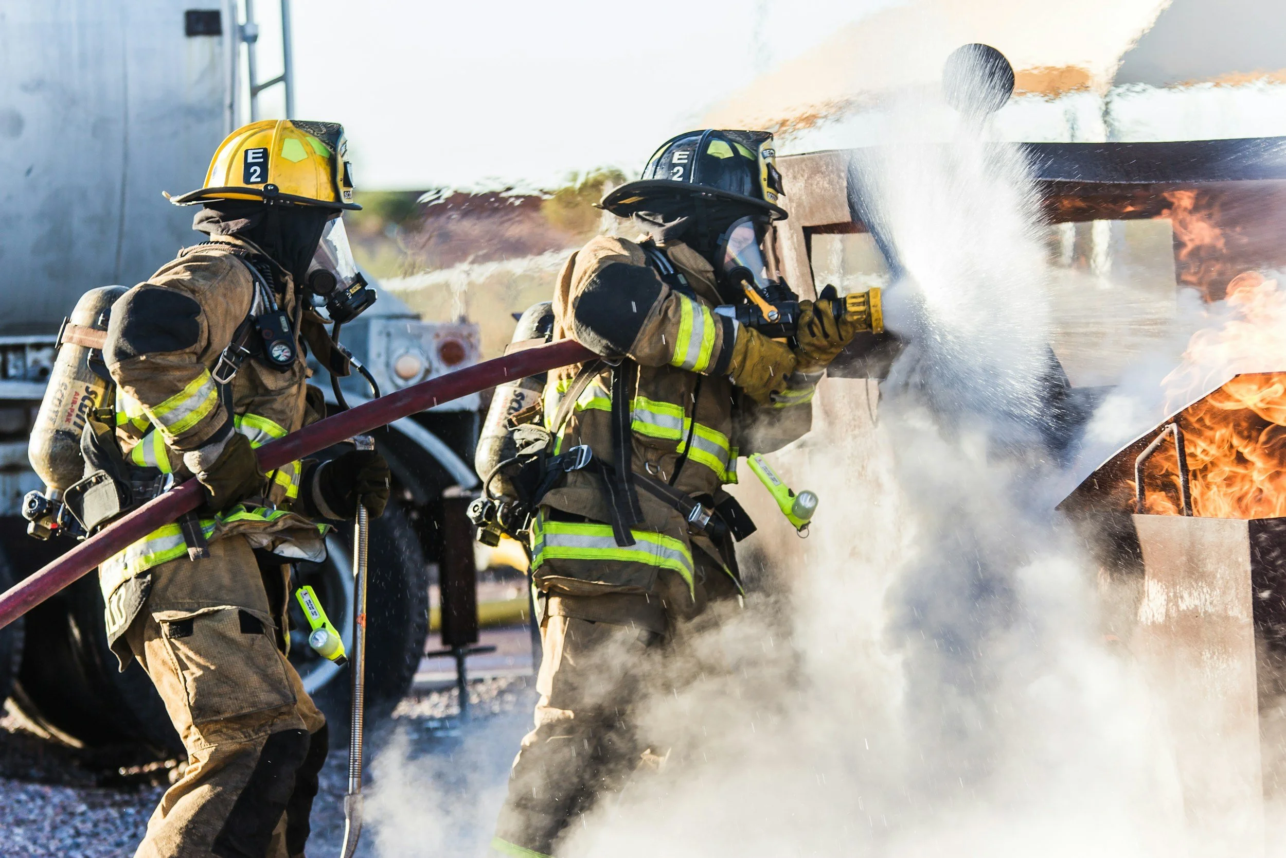 Two firefighters in full gear extinguishing a fire in a vehicle with a hose, surrounded by smoke and flames.