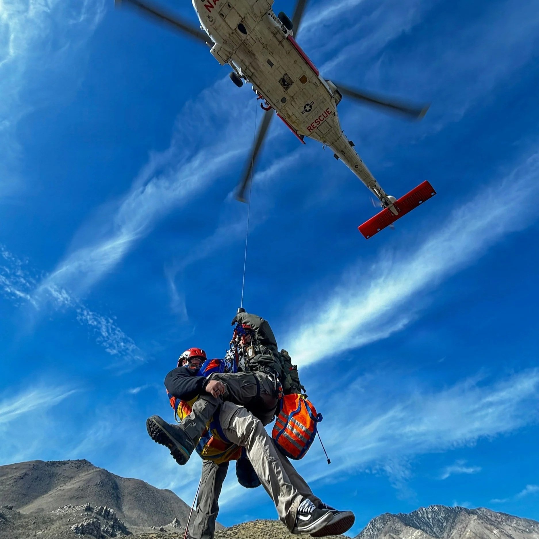 A rescue worker is being airlifted by a helicopter over mountainous terrain, wearing safety gear and a helmet.