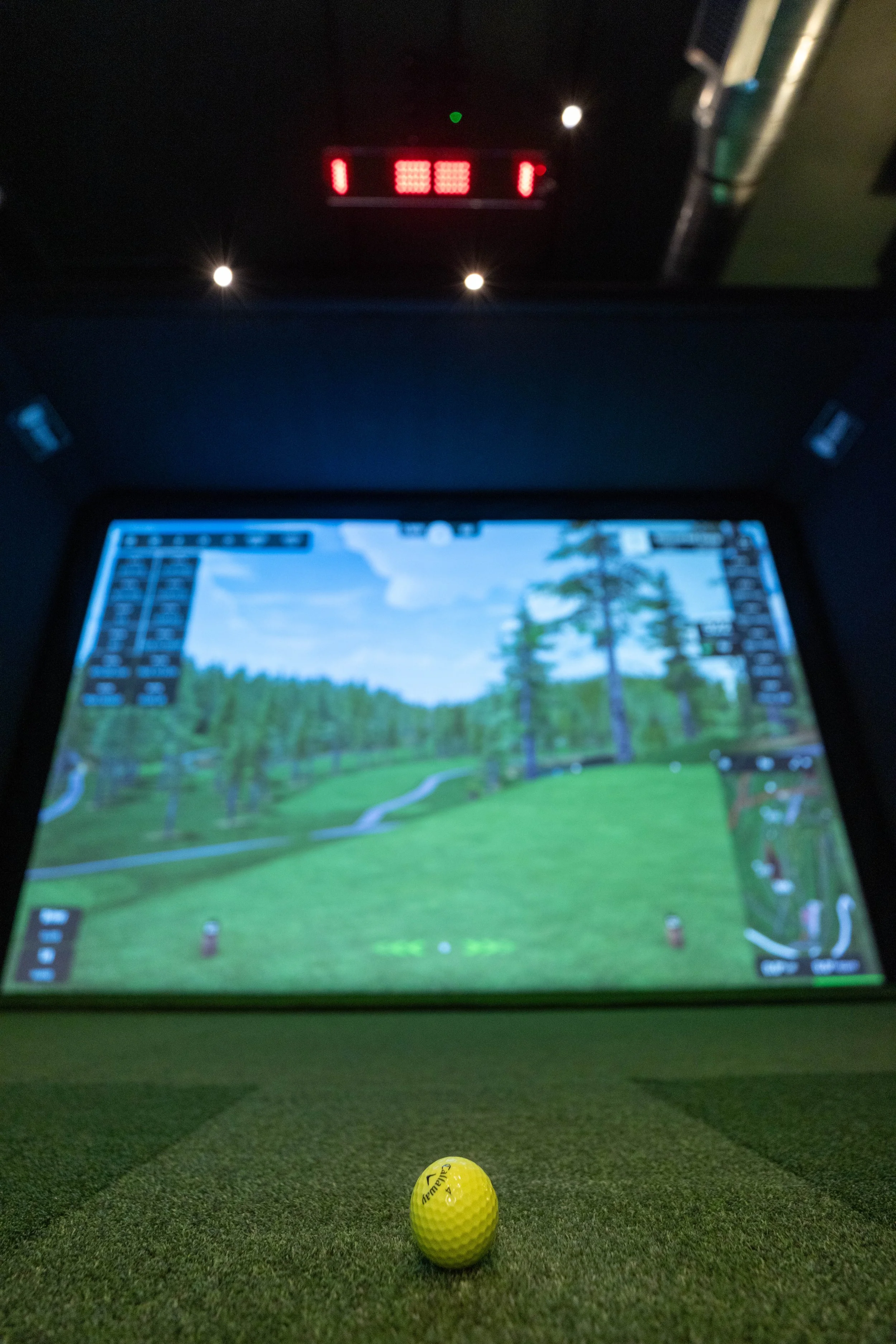 A yellow golf ball positioned on a green indoor golf simulation mat with a digital golf course screen in the background and a red digital score display on the ceiling.