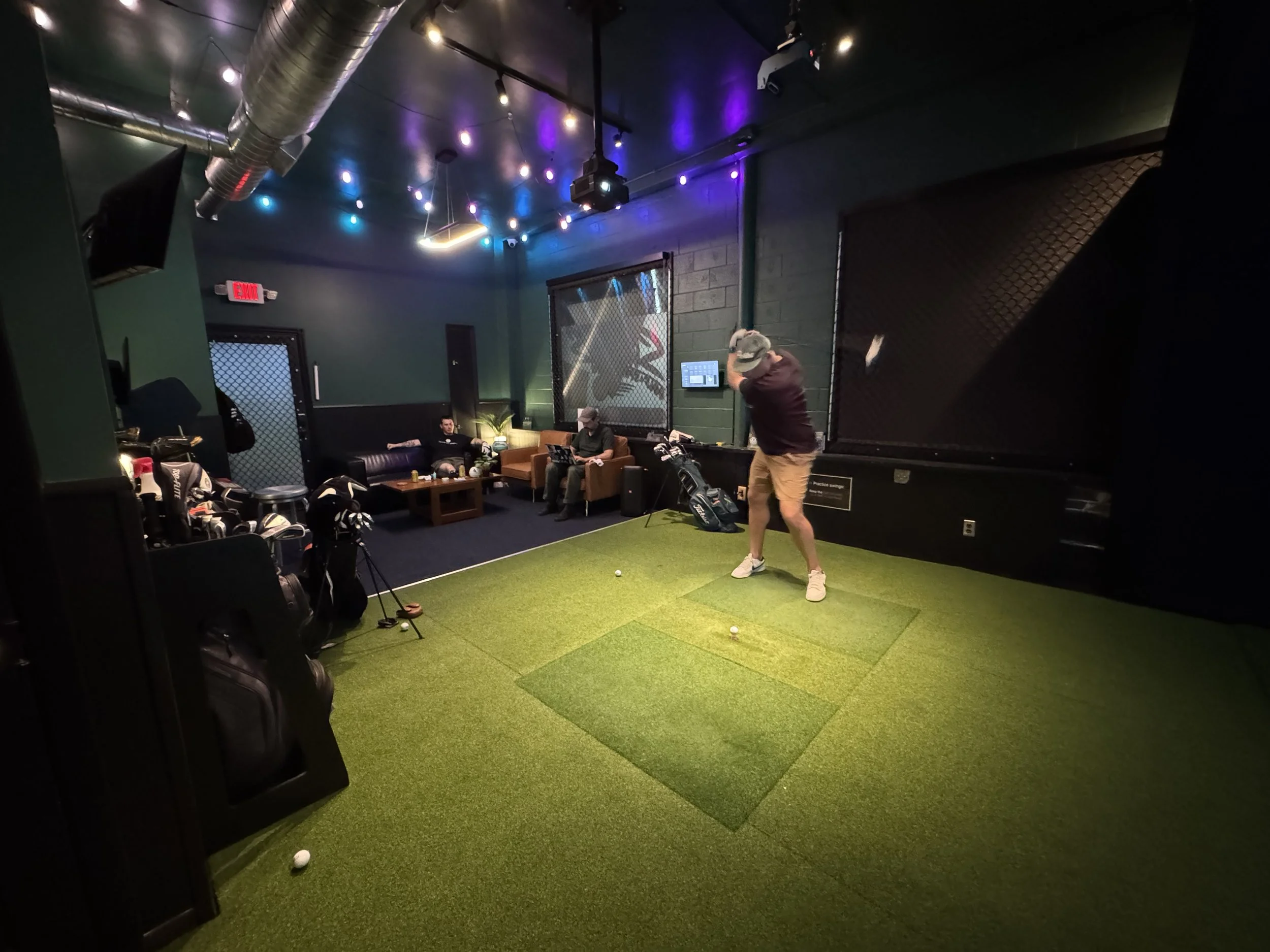 A group of friends relaxes in the seating area while another person plays golf in the simulator at Swingers Golf Club.