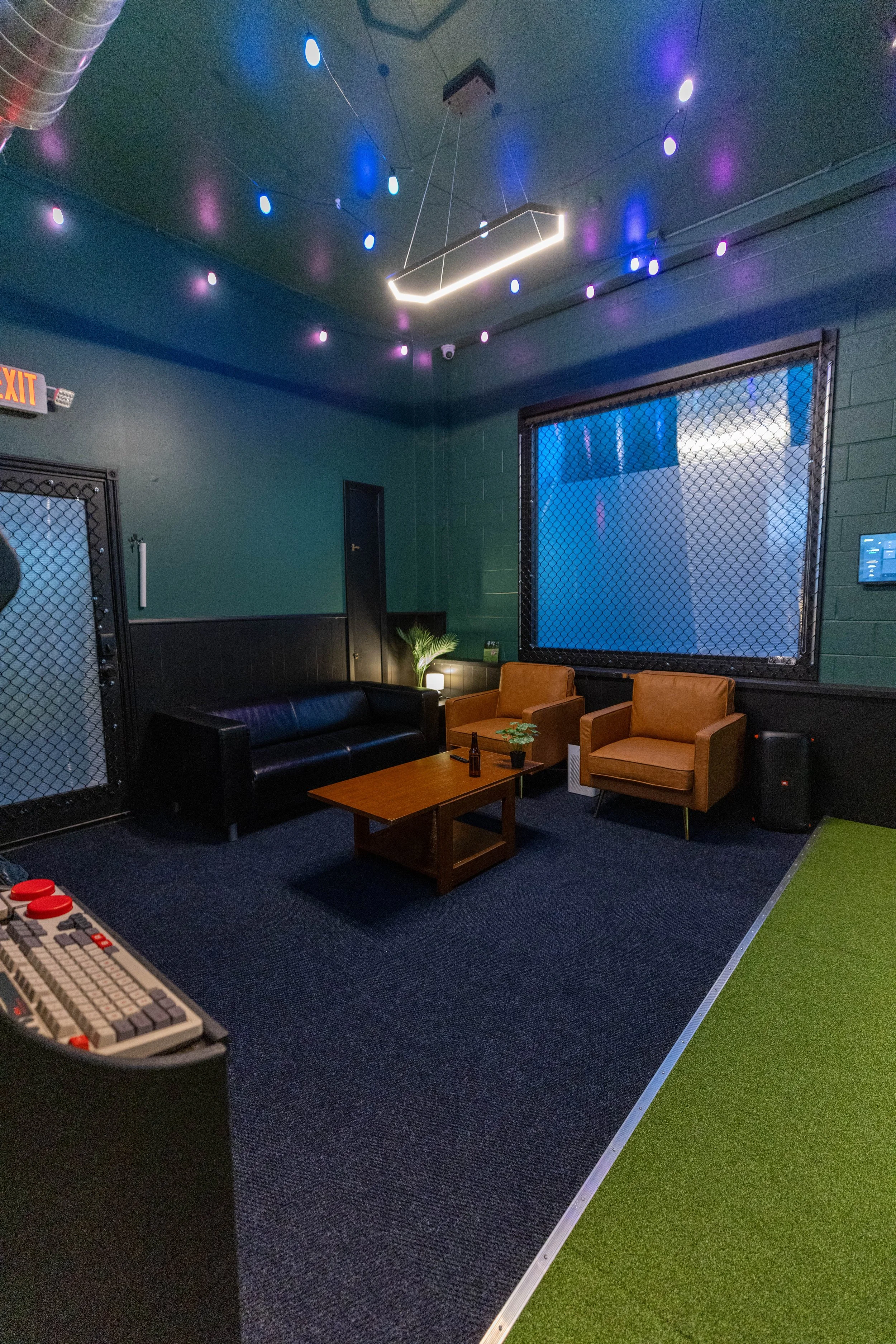 Indoor gaming or entertainment room with dark green walls, black and brown furniture, large window with a wire mesh, and decorative ceiling lights. Part of a gaming console or controller is visible at the bottom left. Carpeted floor with blue and green sections.