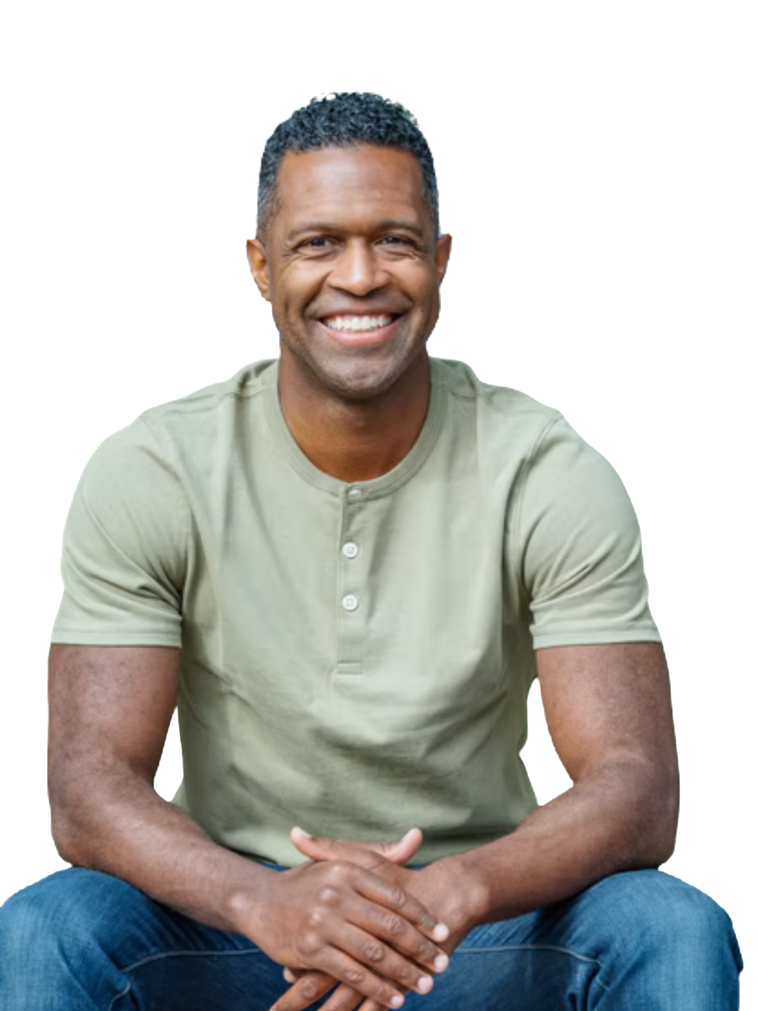 A smiling man with short curly black hair, wearing a light green buttoned shirt and blue jeans, sitting with hands clasped in front of him.