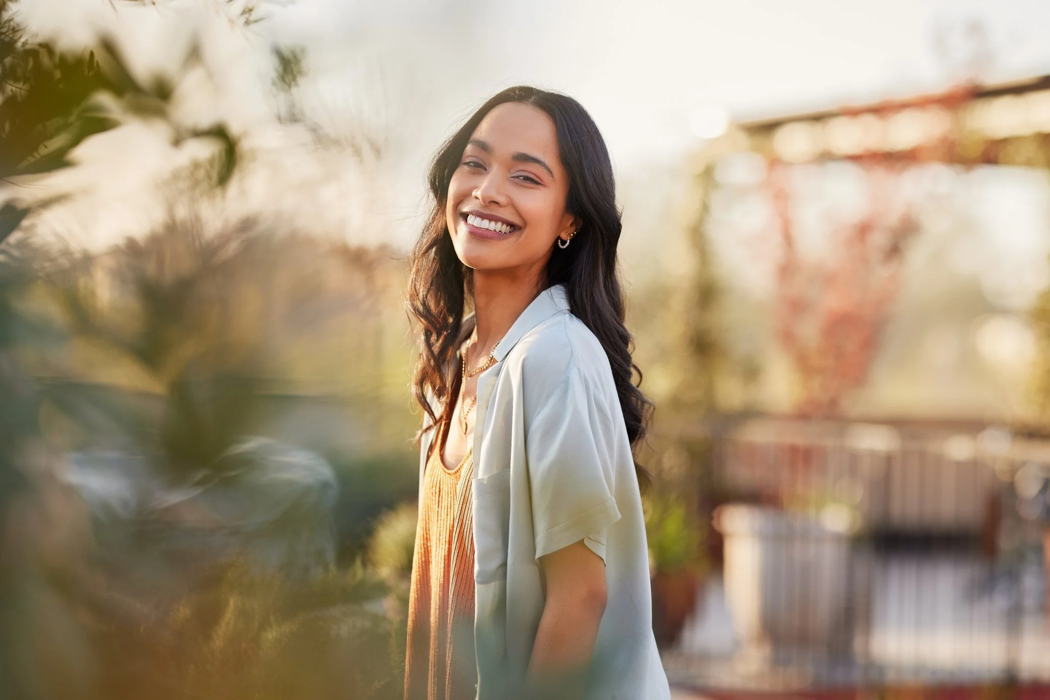 A young woman with dark hair, smiling and looking at the camera, outdoors in sunlight, with trees and a fence in the blurred background.