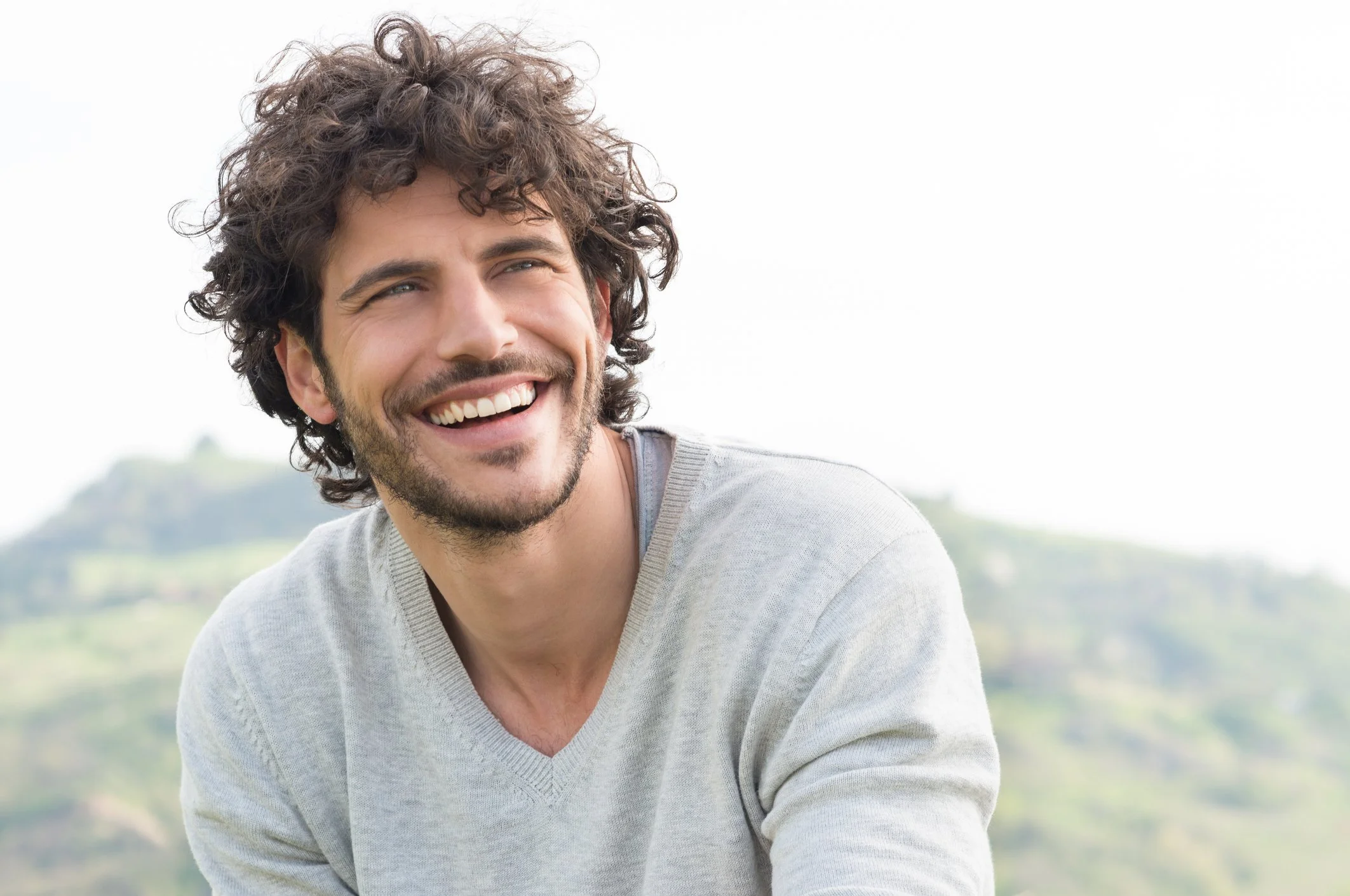 A young man with curly dark hair, a beard, and a mustache smiling outdoors. He is wearing a light gray sweater, against a backdrop of green hills and a partly cloudy sky.